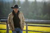 Kevin Costner as John Dutton in Yellowstone, stood in a field next to a fence, his hands in his pockets, wearing a cowboy hat (Credit: Paramount+) 