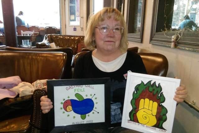 Arlene Wege pictured while being interviewed by the Journal back in 2019 in Derry with a depiction of one of the iconic murals she painted beside Free Derry Corner and her design for the first ever Gasyard Feile, which she hand painted onto T-shirts to sell at the time. Local man Frankie McMenamin had an original T-shirt from the time which helped Arlene to recreate the artwork for the Museum of Free Derry where she volunteered. She had also created new artworks of the Fist in the Flame mural artwork to be sold at the Museum of Free Derry during the 50th anniversary of the Battle of the Bogside.
