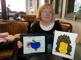 Arlene Wege pictured while being interviewed by the Journal back in 2019 in Derry with a depiction of one of the iconic murals she painted beside Free Derry Corner and her design for the first ever Gasyard Feile, which she hand painted onto T-shirts to sell at the time. Local man Frankie McMenamin had an original T-shirt from the time which helped Arlene to recreate the artwork for the Museum of Free Derry where she volunteered. She had also created new artworks of the Fist in the Flame mural artwork  to be sold at the Museum of Free Derry during the 50th anniversary of the Battle of the Bogside.