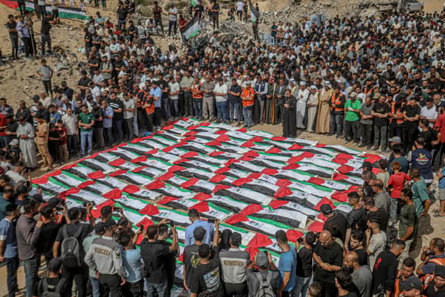 An elevated view shows mourners standing closely around rows of Palestinian flag-draped shrouds during a mass funeral amid rubble in Gaza City, Gaza Strip, on October 1, 2026. Gaza Civil Defense said the remains were among those of 105 people recovered from beneath the rubble of the Al-Taj residential complex and surrounding buildings in the Al-Yarmouk area, which were destroyed in Israeli strikes on October 25, 2023. The remains were taken for burial at Sheikh Radwan cemetery after the funeral. (Photo by Faiz Qreqa / Middle East Images / AFP via Getty Images)